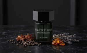 Елегантна продуктова фотография на бутилката Yves Saint Laurent La Nuit de L’Homme Le Parfum, поставена на тъмна повърхност. Около основата на бутилката са подредени ключовите ароматни съставки: зърна черен пипер, звездовиден анасон, сушени стръкове лавандула и парчета кехлибар. Светлината е мека и подчертава детайлите, на тъмен, леко разфокусиран заден план